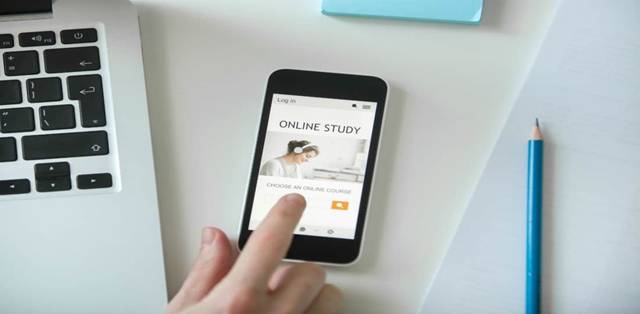 A Human Hand Touches the SmartPhone Which Showing Online Study Application That Placed On The Table Near The Laptop.