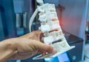 Close up mage of a persons hand holding a white 3D model of a portion of human spine inside a blurred printer room.