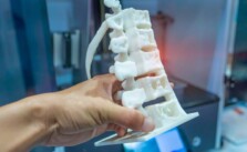 Close up mage of a persons hand holding a white 3D model of a portion of human spine inside a blurred printer room.