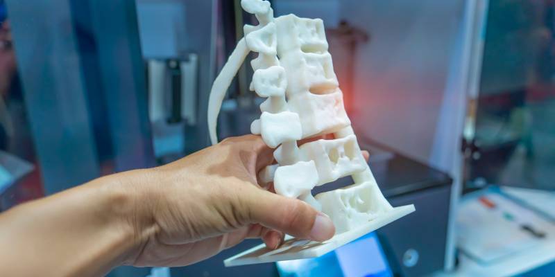 Close up mage of a persons hand holding a white 3D model of a portion of human spine inside a blurred printer room.